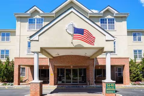 Three-story beige senior living building with brick entrance and waving American flag.