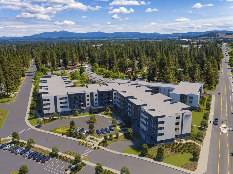Aerial view of Rockwood at Whitworth, a large multi-story senior living facility surrounded by trees and greenery. The building has a modern design with multiple wings and a parking lot with several cars in front. The facility is located near a road with light traffic and is set against a backdrop of forested hills and a partly cloudy sky.