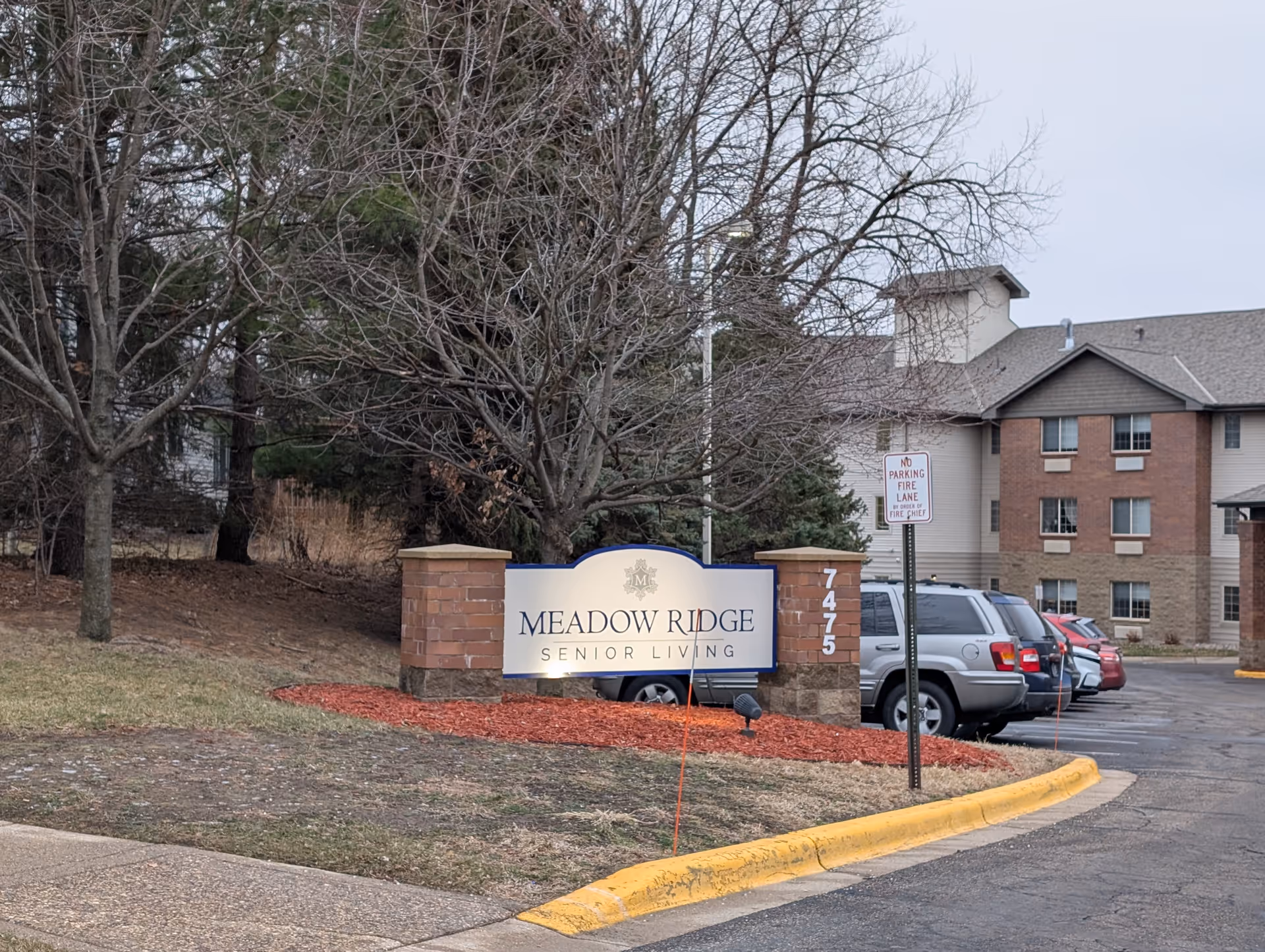 Stone-and-brick entrance sign reading 'Meadow Ridge Senior Living' in front of a multi-story senior living building with parked cars and leafless trees.