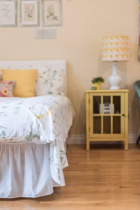 Cozy bedroom with a floral bedspread, yellow pillows, and a yellow nightstand with a lamp.