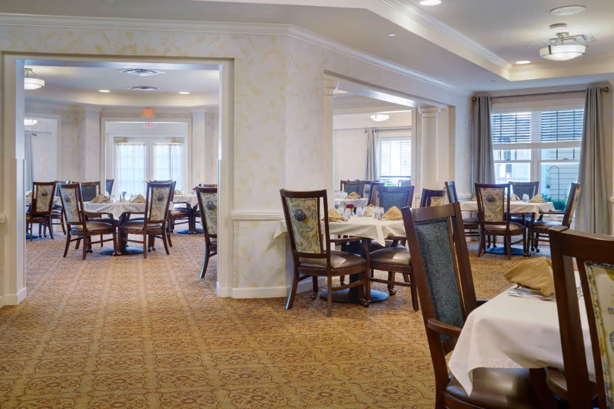 Interior view of a senior living facility dining room with multiple round tables covered with white tablecloths, each set with napkins, glasses, and silverware. The room has patterned carpet, floral upholstered chairs, large windows with curtains, and soft ceiling lighting.
