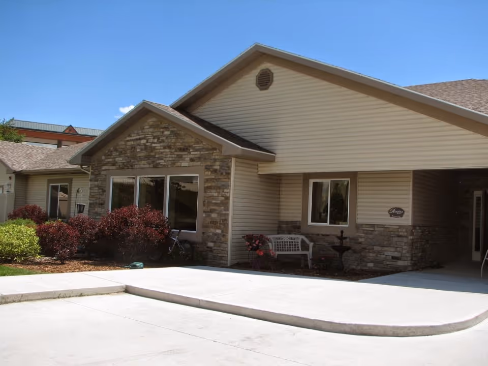 Front exterior of a single-story assisted living building with stone and siding facade, shrubs, bench, and a curved driveway.