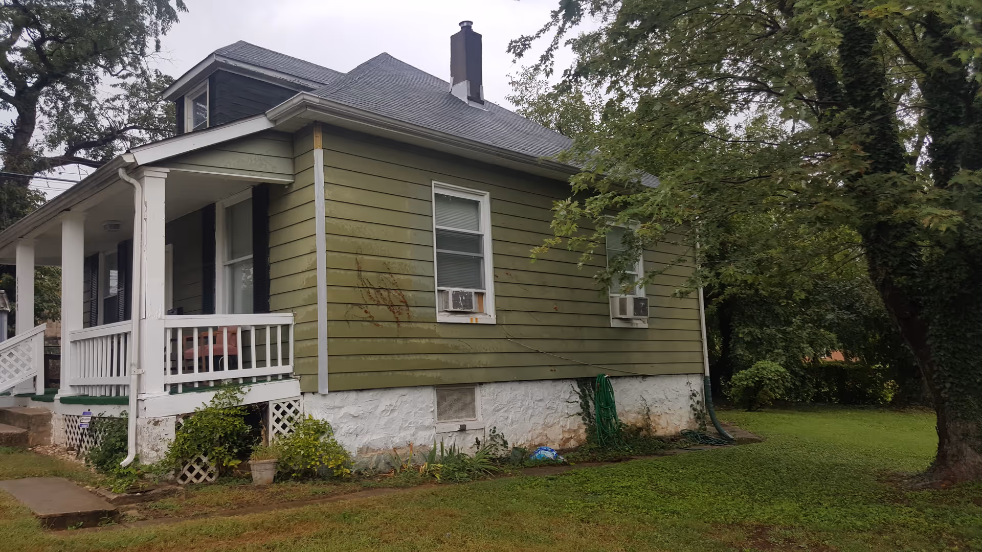Exterior view of a single-story green house with white trim and a small porch. The house has two windows with air conditioning units and a chimney on the roof. There is a garden hose coiled near the foundation and trees surrounding the property.