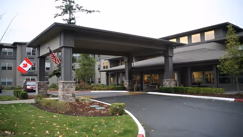 Entrance of Port Townsend Senior Living by Cogir featuring a covered driveway supported by stone and dark pillars, with the American and Canadian flags displayed on the left side. The building is a multi-story structure with large windows and landscaped greenery around the entrance.