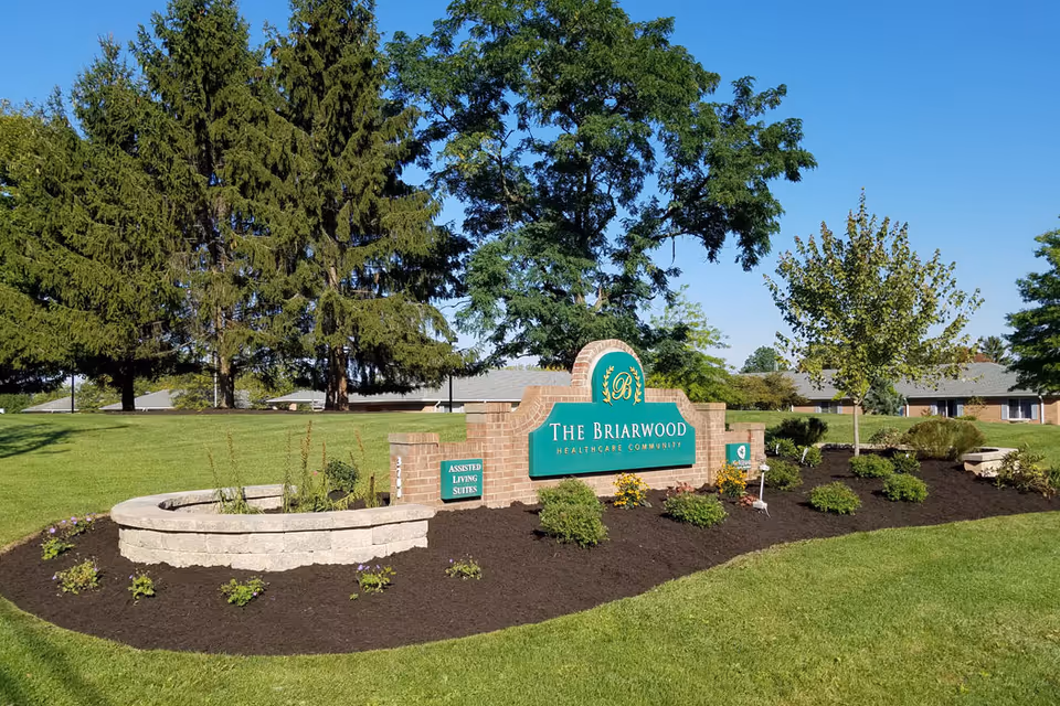 A landscaped outdoor area with a brick sign that reads 'The Briarwood Healthcare Community' surrounded by small bushes, flowers, and trees under a clear blue sky.