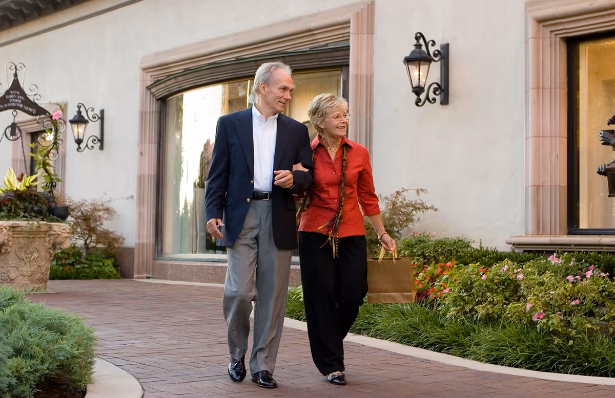 An elderly couple walking arm in arm along a paved pathway outside a building with large windows and decorative wall lamps. The woman is carrying a brown paper shopping bag and both are smiling, surrounded by greenery and flowers.