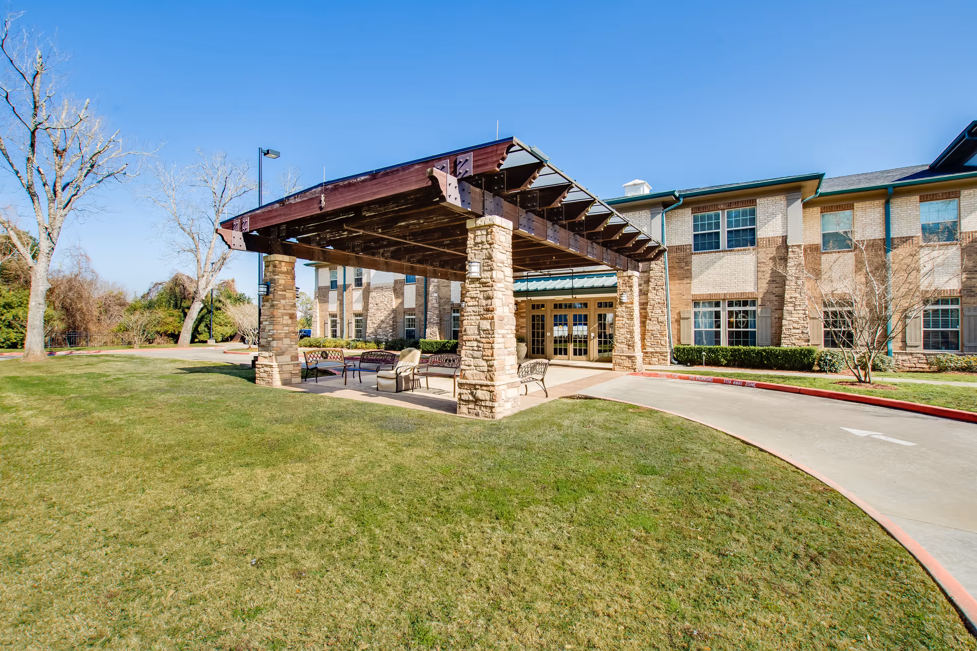 Front entrance of a senior living building with a covered porte-cochere, outdoor seating, and a grassy lawn.