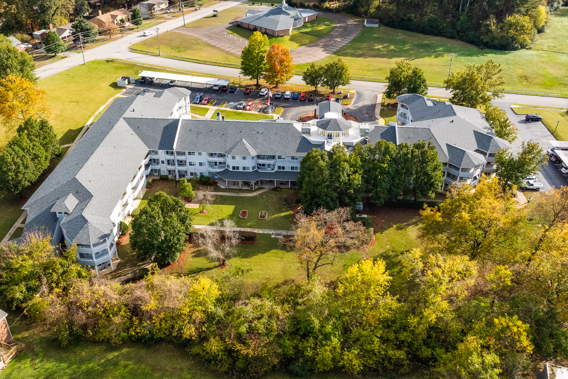 Aerial view of Jackson Meadow, a large senior living community building surrounded by green lawns, trees, and parking areas. The building has multiple wings and a gray roof, with a circular driveway and several parked cars visible.