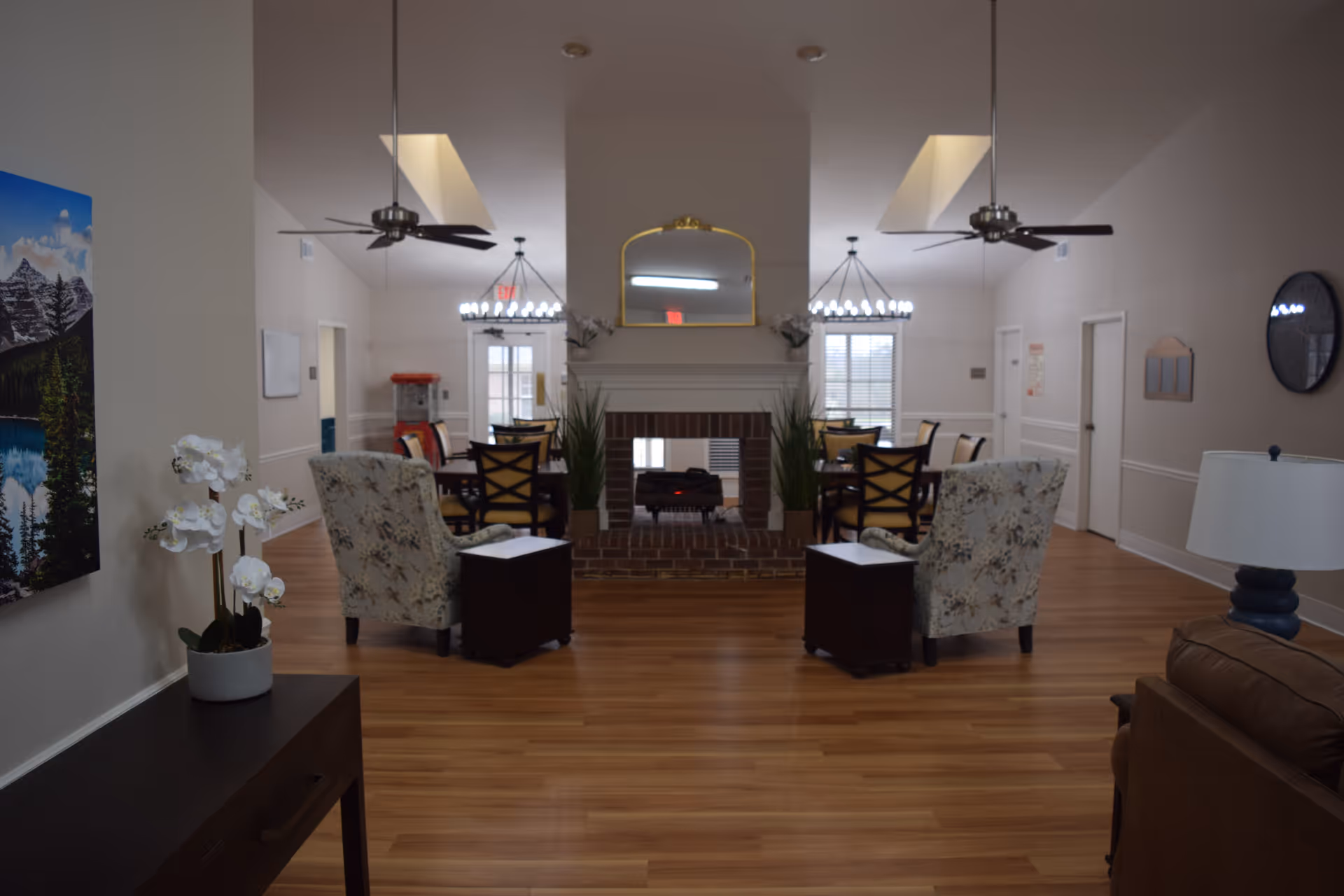Spacious communal living area with chairs and small tables facing a central brick fireplace, dining tables in the background, ceiling fans, and wood flooring.