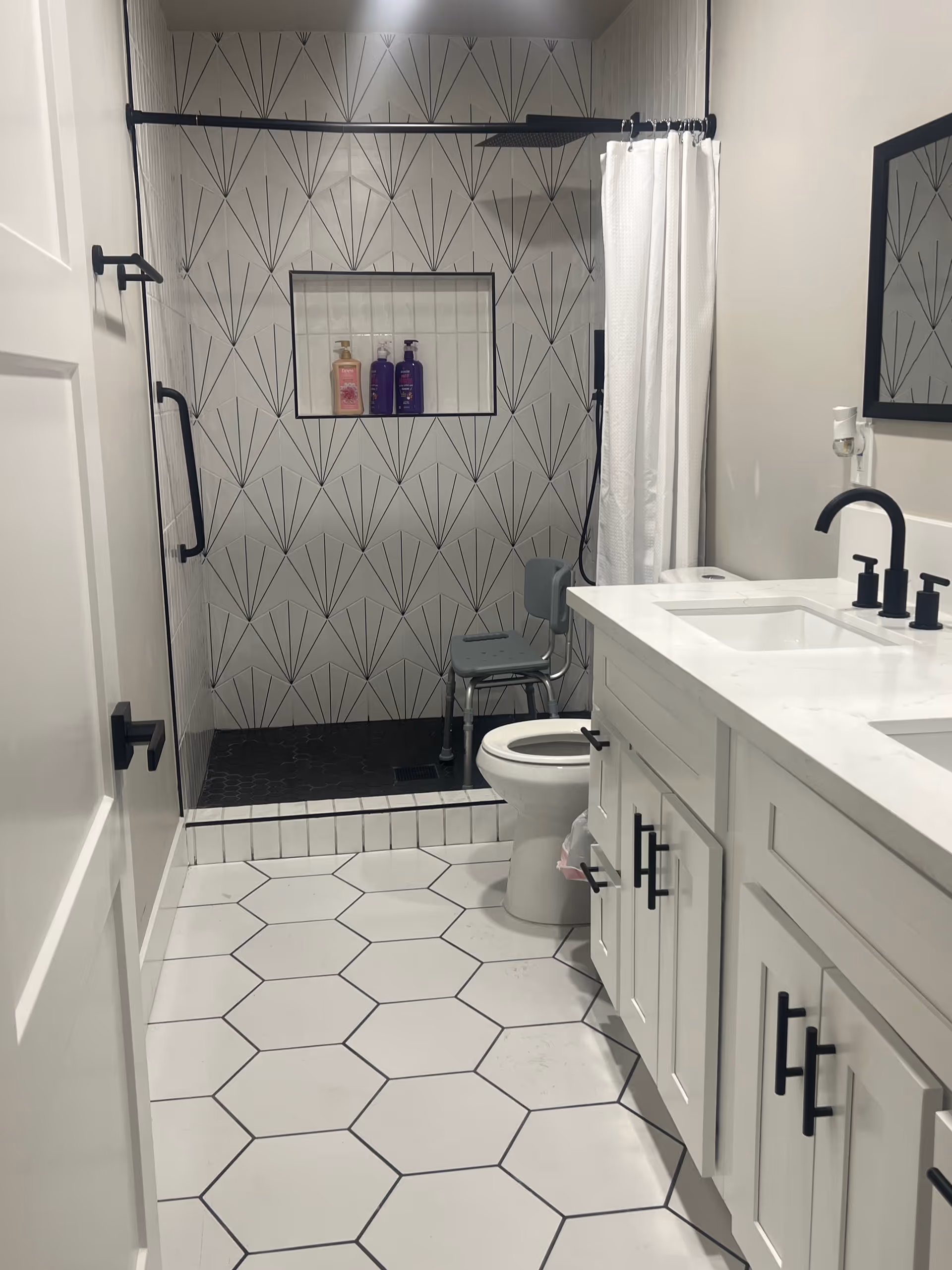 Bright modern bathroom featuring a walk-in shower with patterned tile and a shower chair, white hexagon floor tiles, toilet, and a white double-sink vanity.