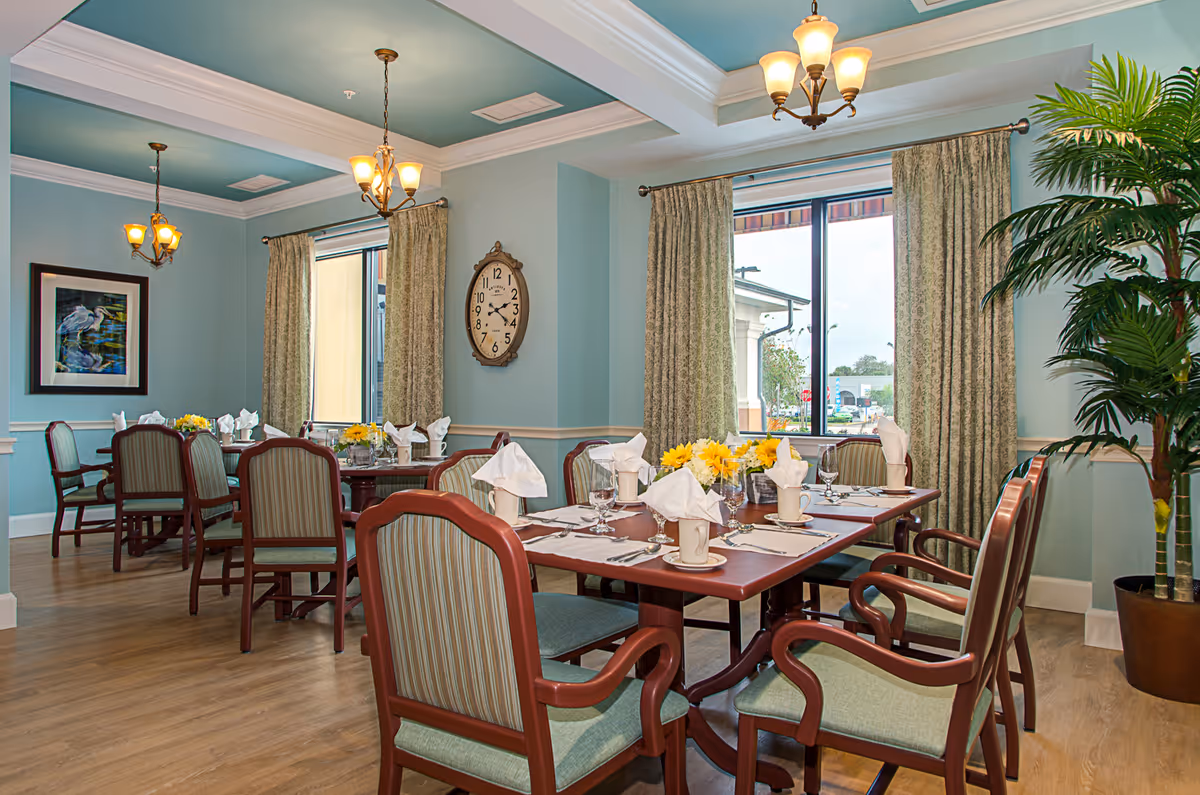 A bright and elegant dining room with wooden tables set for a meal, including white napkins, cups, glasses, and yellow flower centerpieces. The room features light blue walls, large windows with patterned curtains, a large clock on the wall, framed artwork, hanging light fixtures, and a potted plant in the corner.