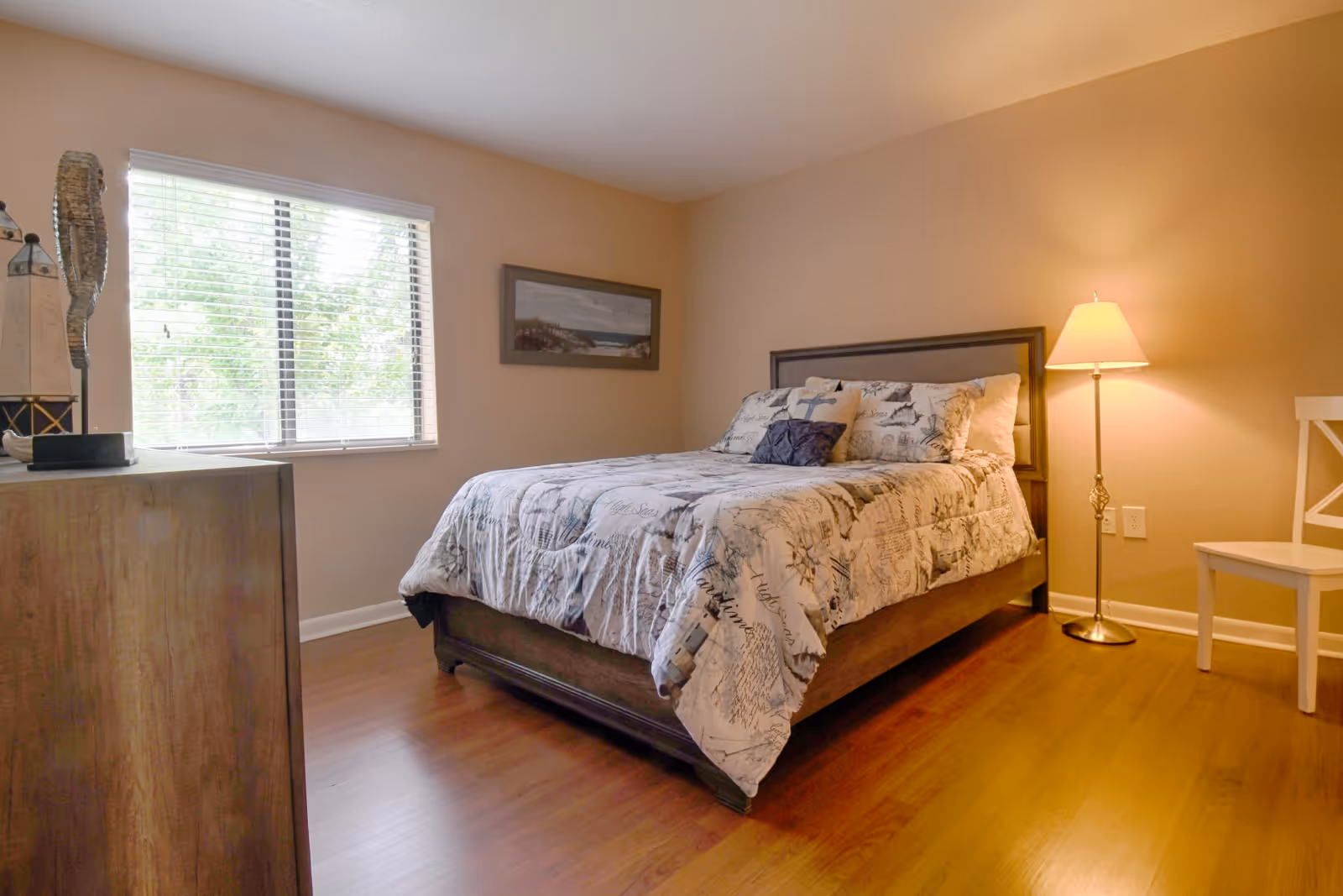A cozy bedroom with a wooden bed frame and a bed dressed in a patterned comforter and pillows. There is a wooden dresser on the left side with decorative items, a window with blinds letting in natural light, a framed picture on the wall, a floor lamp emitting warm light, and a white chair on the right side. The room has wooden flooring and beige walls.