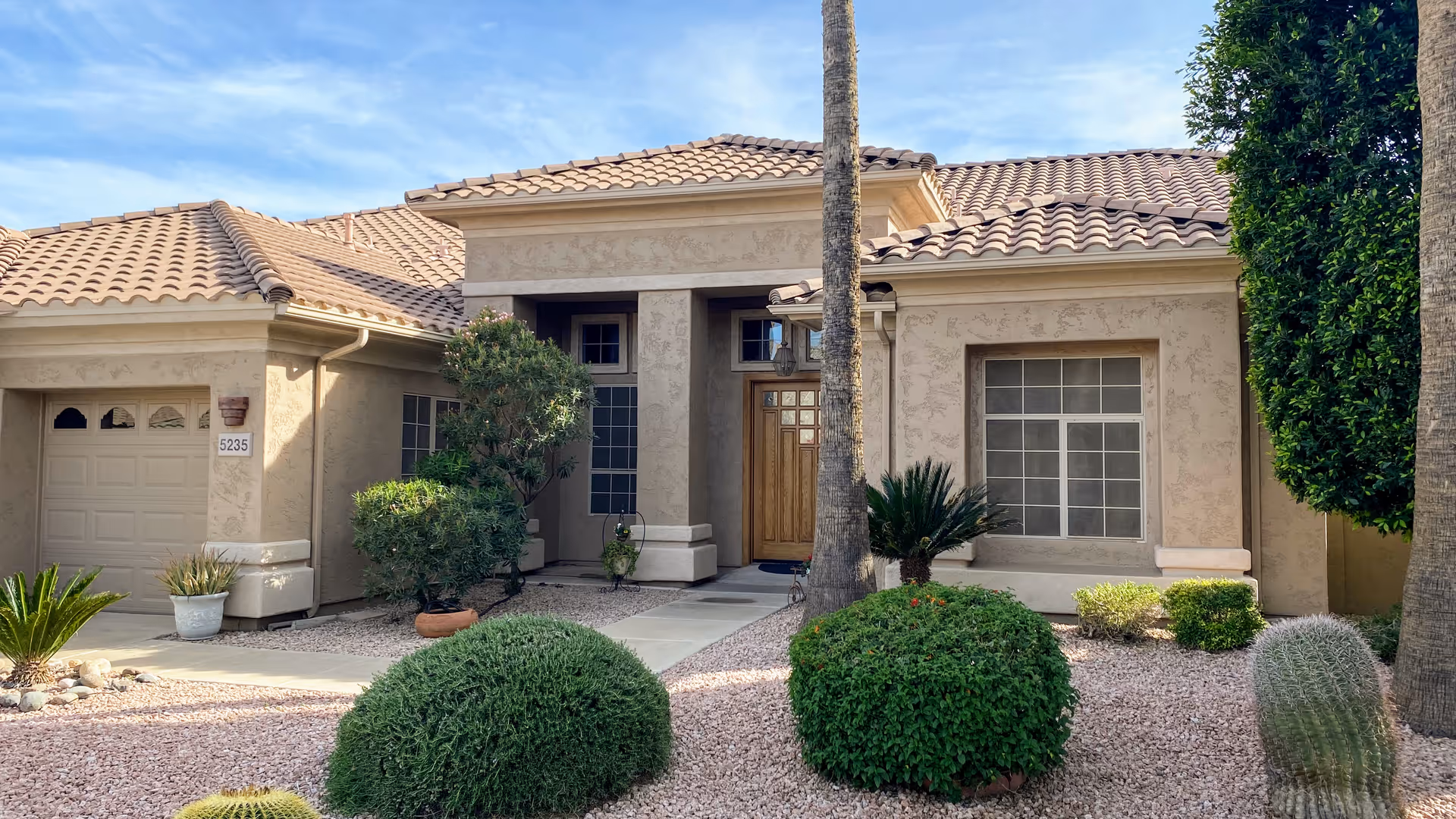 Front exterior view of a single-story residential building with a tiled roof, a wooden front door, large windows, and a garage. The front yard features desert landscaping with gravel, bushes, small trees, and palm trees under a partly cloudy sky.
