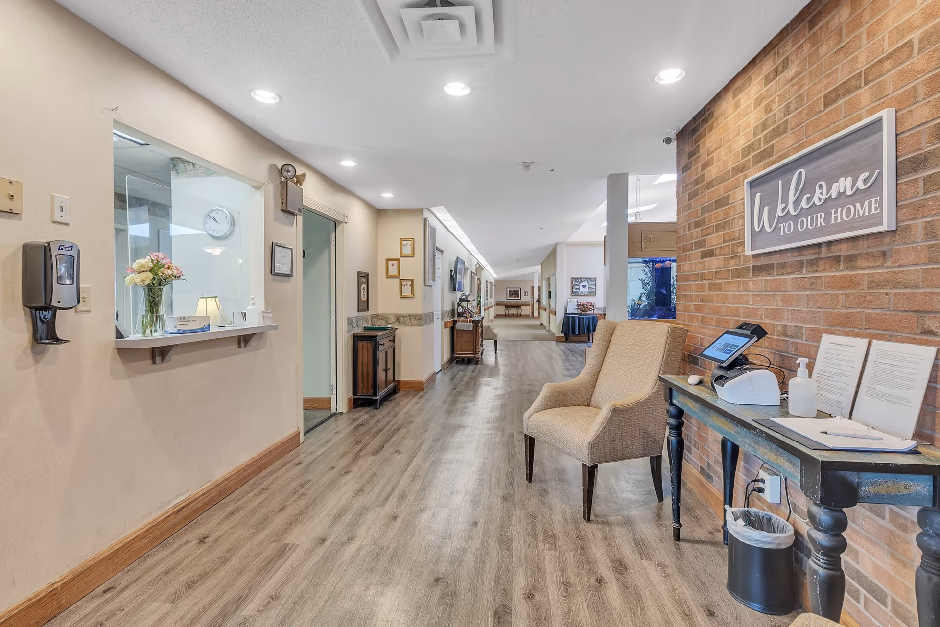 A clean and well-lit hallway in a senior living facility with wood flooring, beige walls, and a brick accent wall. On the right side, there is a small table with a chair, a hand sanitizer dispenser, a sign that reads 'Welcome to Our Home,' and some documents. On the left side, there is a reception window with a vase of flowers and a hand sanitizer dispenser mounted on the wall. The hallway extends into the distance with framed pictures and furniture along the walls.
