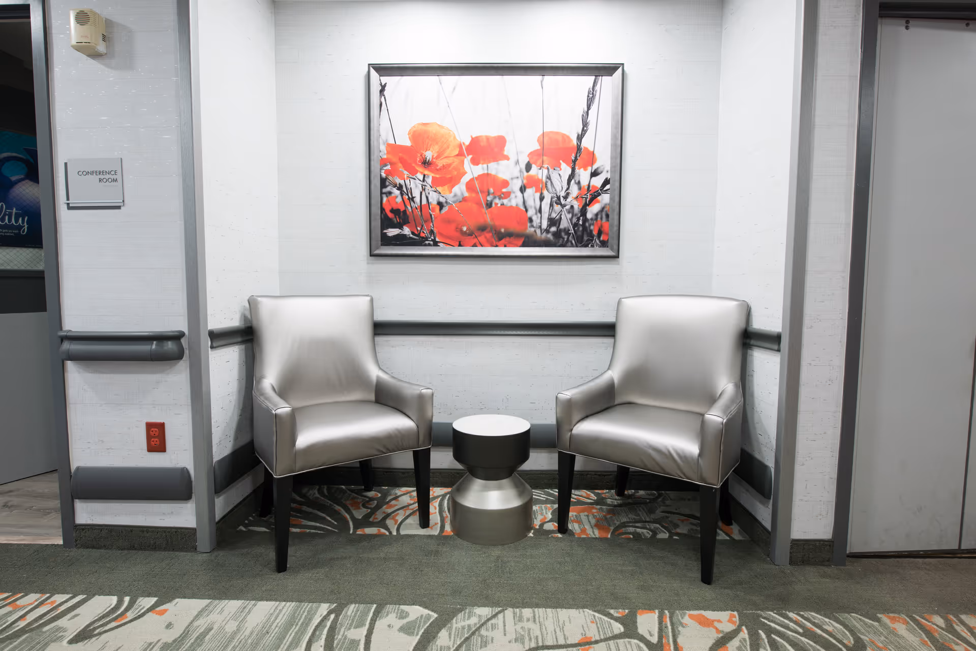 A small seating area with two silver armchairs and a small round silver table between them. Above the chairs is a framed picture of red flowers. The walls are light-colored with gray trim, and there is a sign on the left wall indicating a conference room.