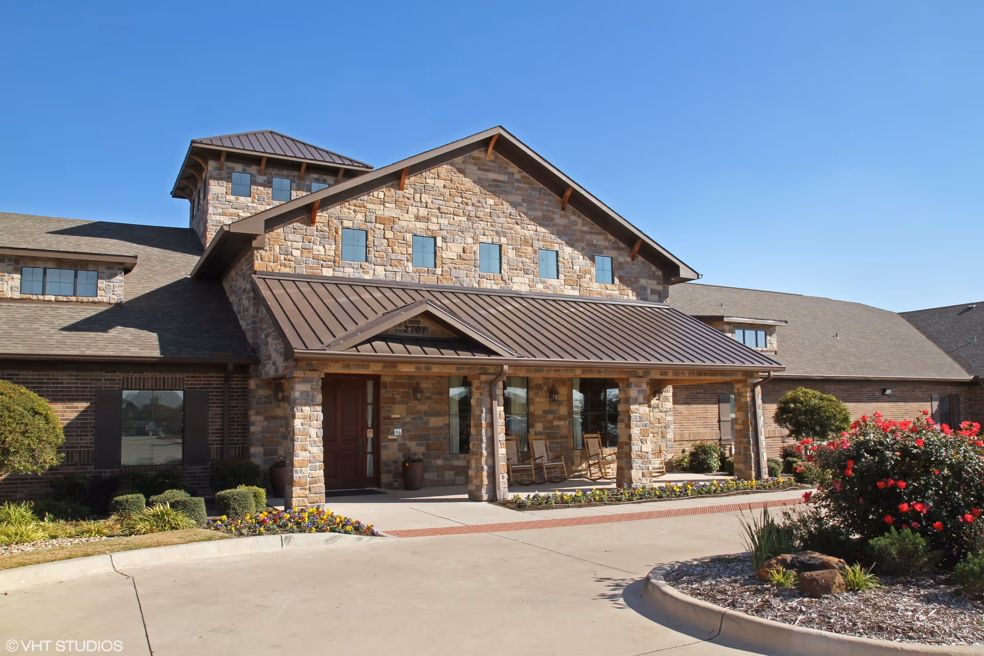 Stone building front with a covered porch, rocking chairs, stone facade and landscaped driveway under a clear blue sky.