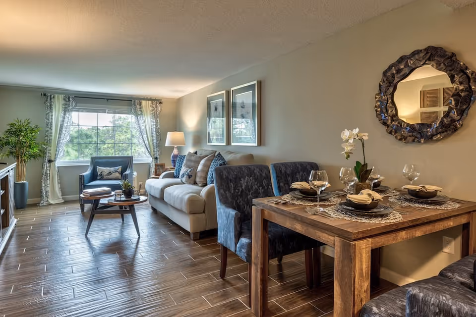 A cozy living and dining area with a beige sofa, blue armchair, wooden coffee table, and a wooden dining table set for four with plates, bowls, napkins, and wine glasses. The room features a large window with sheer curtains, a decorative round mirror on the wall, two framed pictures, a potted plant, and a table lamp.