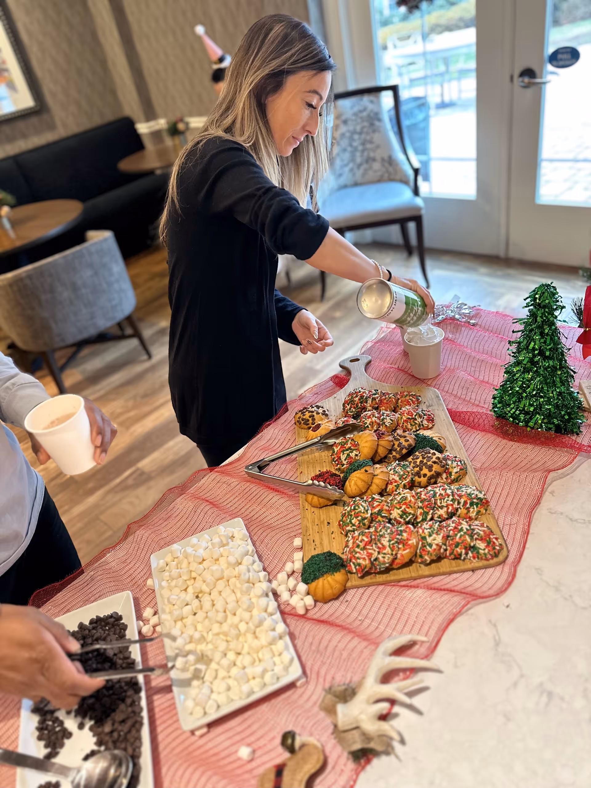A woman pours whipped cream into a cup at a festive dessert and hot chocolate station with cookies, marshmallows, and chocolate chips on a table in a communal dining area.
