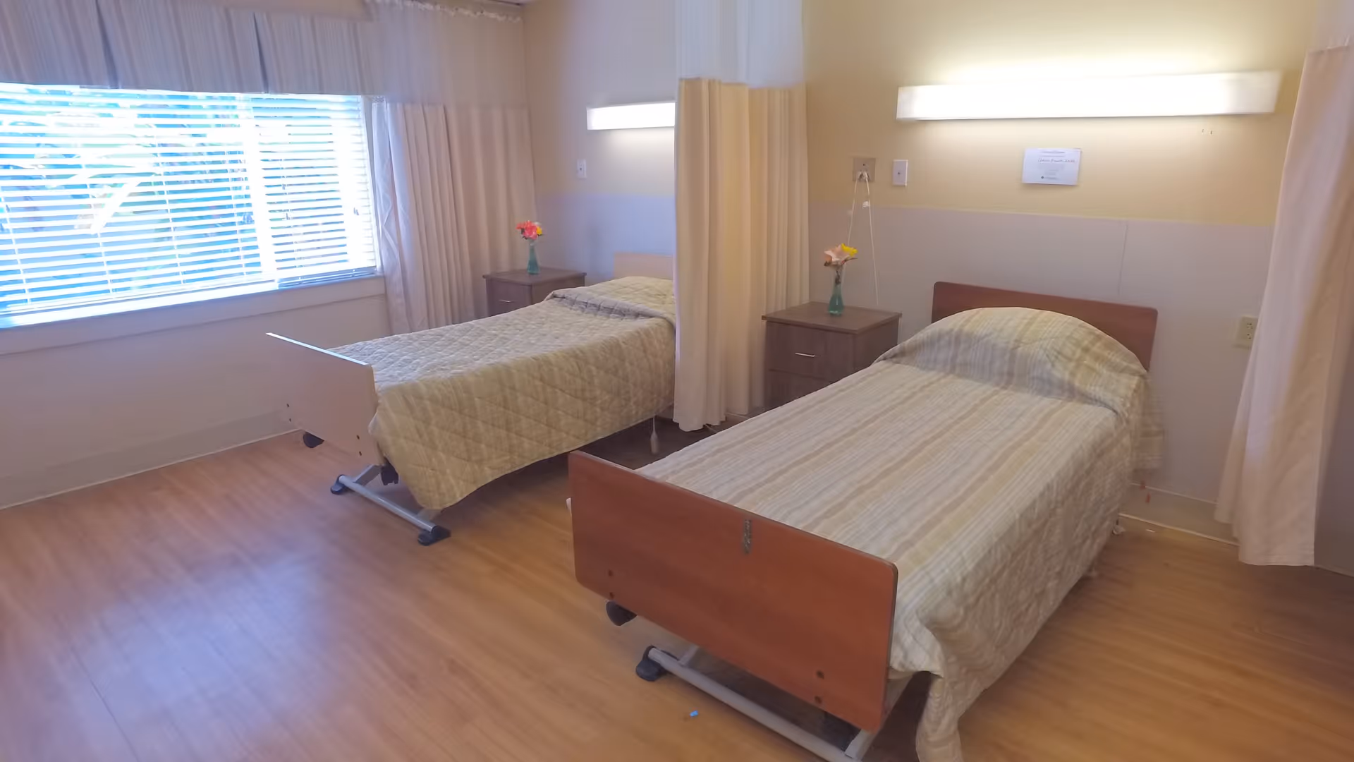 A room with two single beds separated by a curtain. Each bed has a wooden headboard and footboard, and is covered with a light-colored quilt. There are two small bedside tables, each with a small vase holding flowers. A large window with blinds and curtains lets in natural light, and the floor is wooden.