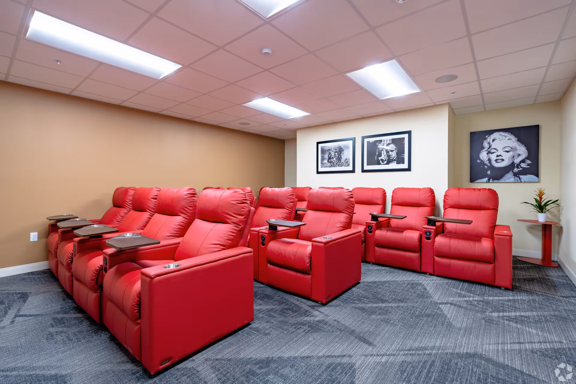 A small theater room with two rows of red leather recliner chairs, each equipped with a wooden tray and cup holder. The walls are beige and cream, decorated with black and white framed pictures including a portrait of Marilyn Monroe. The room has a gray carpet and a drop ceiling with fluorescent lights.