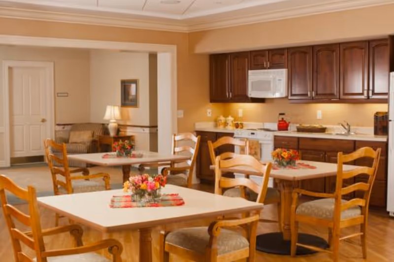 Communal dining area with wooden tables and chairs, floral centerpieces, and a kitchenette with dark cabinets and a microwave.