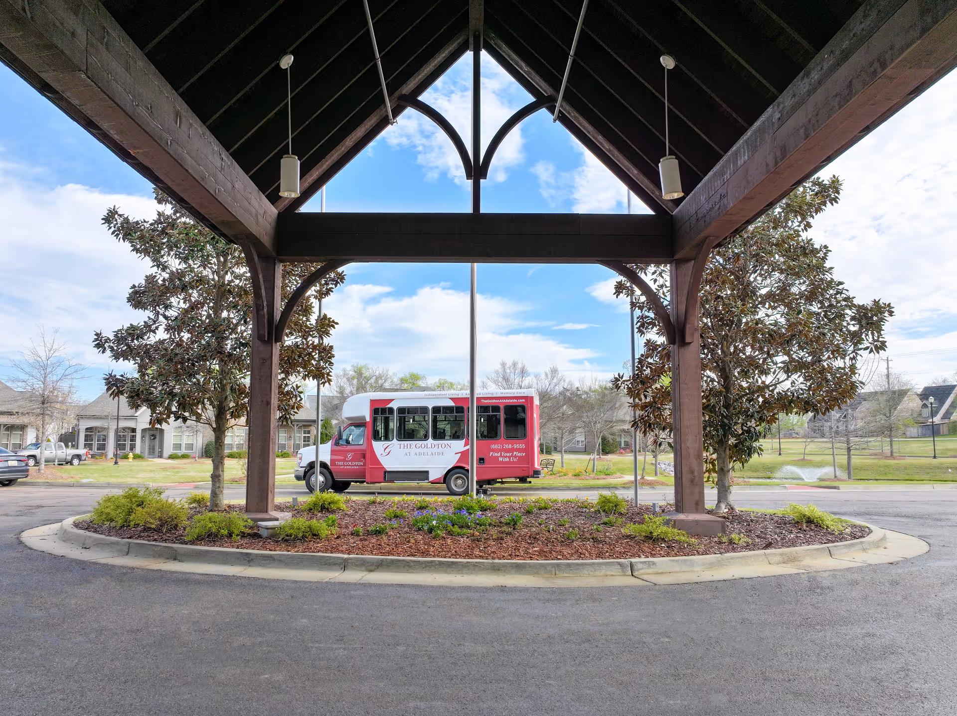View from under a wooden covered driveway entrance looking out towards a small shuttle bus parked in front of a landscaped roundabout with trees and shrubs. The bus has signage for The Goldton at Adelaide senior living facility. Residential buildings and a fountain in a pond are visible in the background under a partly cloudy sky.