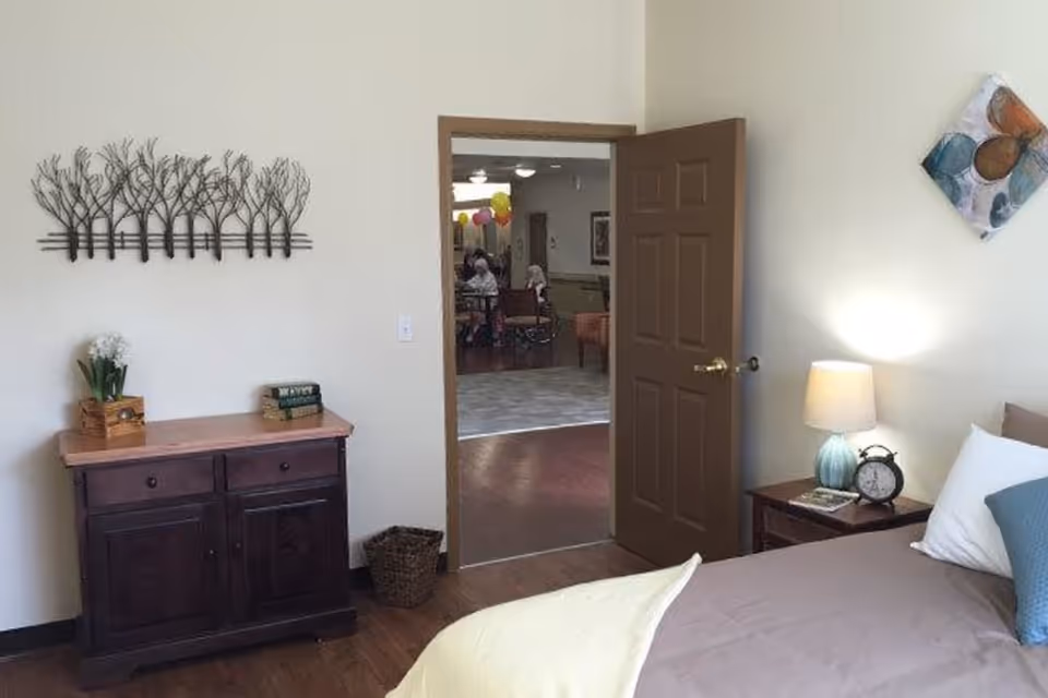 A furnished bedroom with a bed, bedside table and lamp, wooden dresser, and an open door leading to a common area.