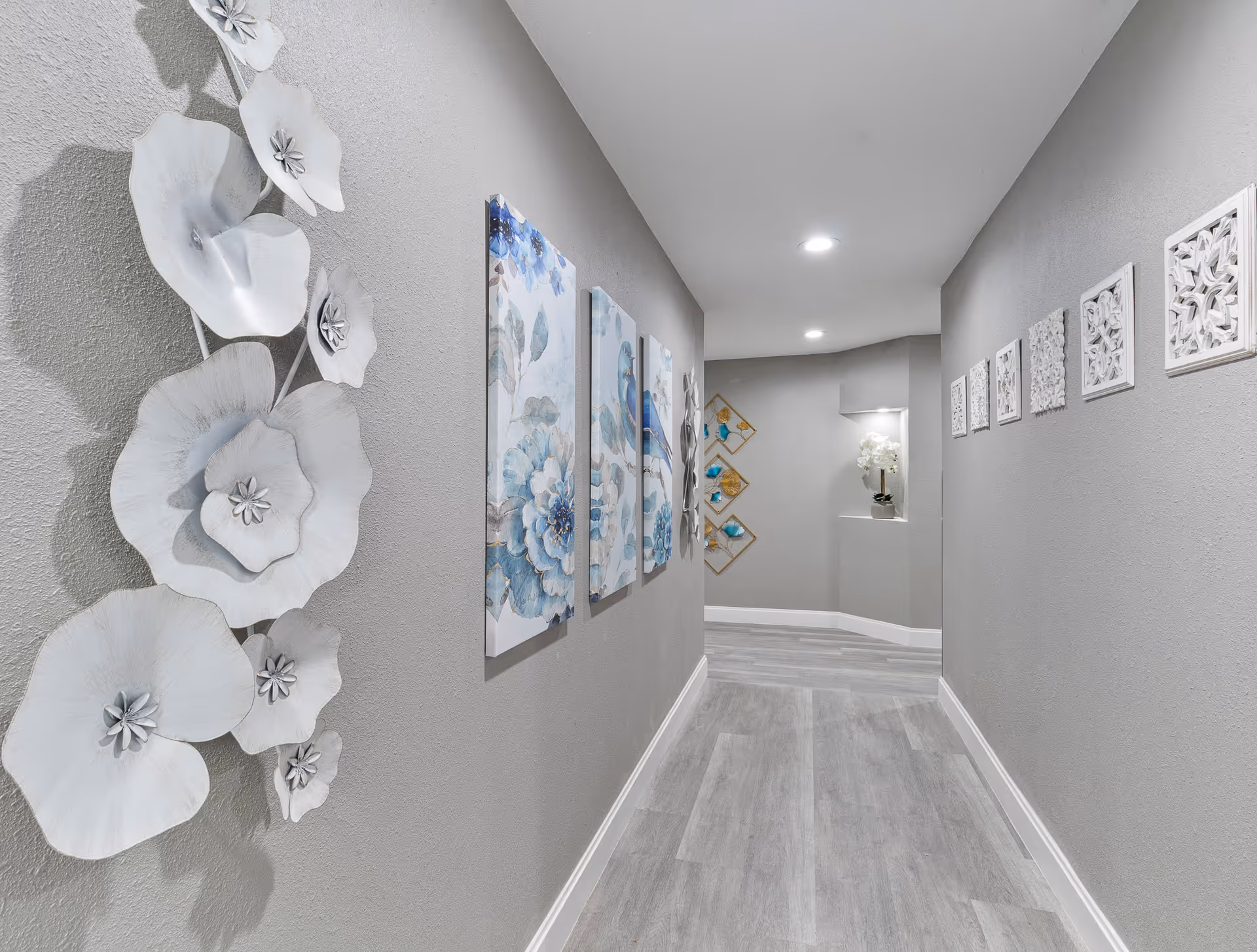 A well-lit hallway with gray walls and light wood flooring. The left wall is decorated with large white metal flower sculptures and three blue floral paintings. The right wall features a series of small white decorative square panels. At the end of the hallway, there is a small alcove with a white flower arrangement and additional wall art with blue and gold accents.
