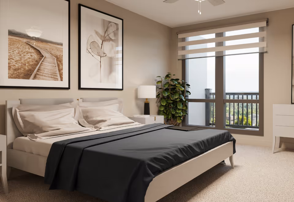 Sunlit bedroom with a made bed, framed wall art, nightstand and lamp, potted plant, and large window overlooking a balcony.