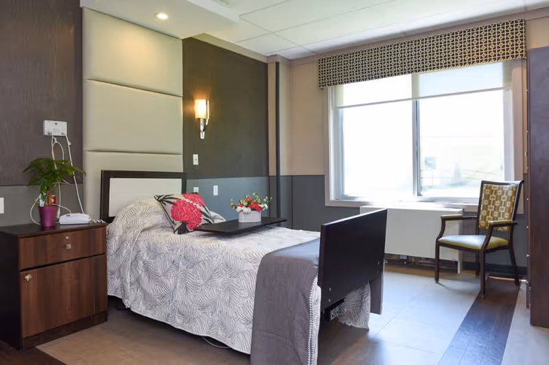 A neatly made single bed in a nursing facility room with a white and gray patterned bedspread, a pink floral pillow, and a tray with a flower arrangement on the bed. Next to the bed is a wooden nightstand with a potted plant and a telephone. The room has a large window with a patterned valance, a green cushioned chair, and soft lighting on the wall.