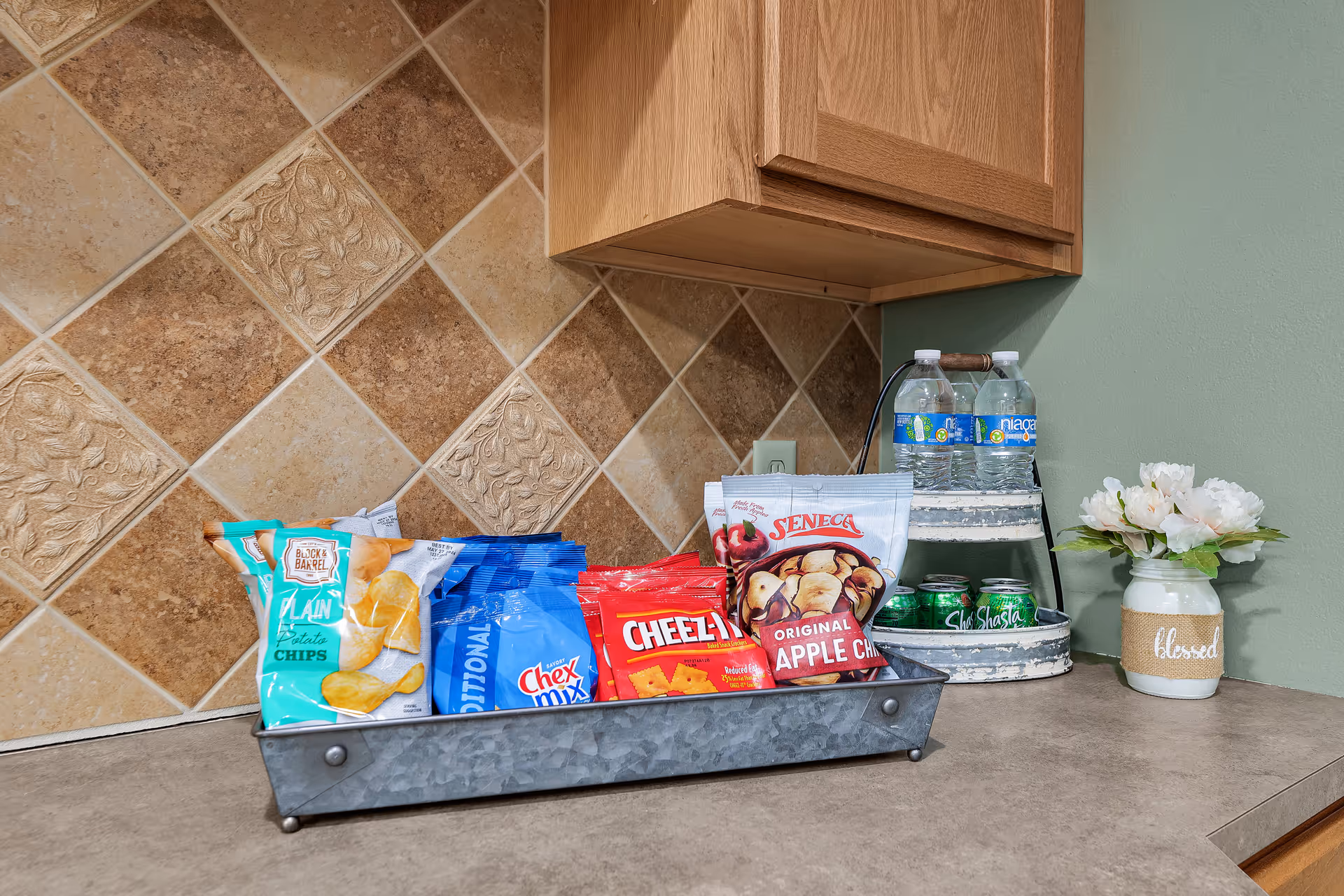A kitchen countertop with a metal tray holding various snack bags including potato chips, Chex Mix, Cheez-It crackers, and Seneca original apple chips. Next to the tray is a two-tiered stand with bottled water and cans of Shasta soda. A small vase with white flowers and a burlap wrap labeled 'blessed' is also on the counter. The backsplash features beige and brown decorative tiles, and there is a wooden cabinet above the counter.