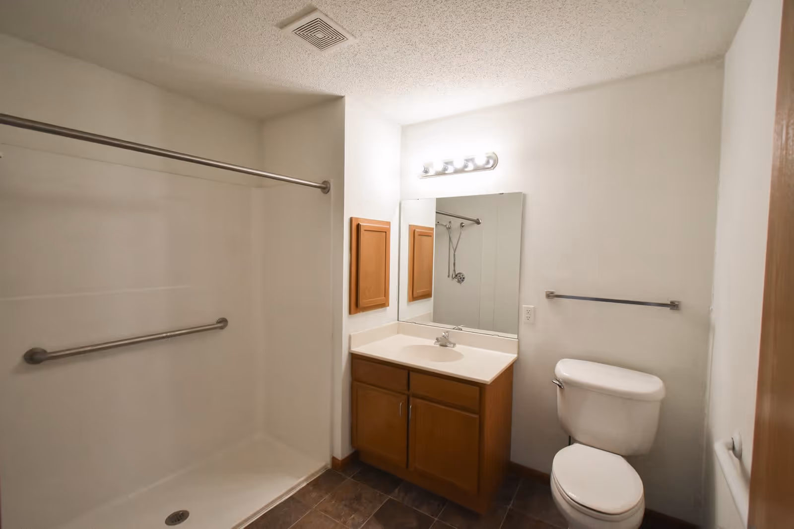A clean bathroom with a walk-in shower featuring a grab bar, a wooden vanity with a sink and mirror above it, a toilet, and a towel rack on the wall. The walls are white and the floor has dark tiles.