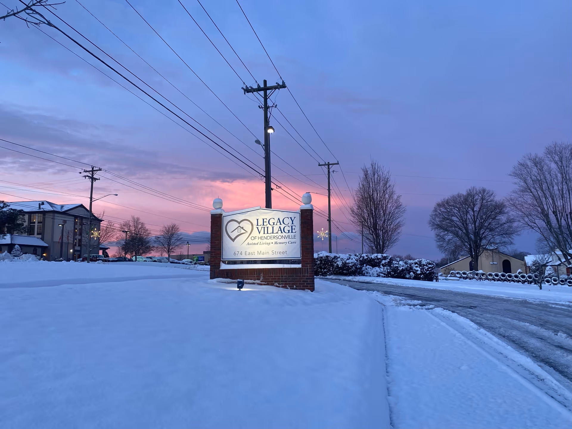 Snow-covered ground and road at dusk with a lit sign for Legacy Village of Hendersonville, an assisted living and memory care facility, located at 674 East Main Street. The sky is a mix of purple and pink hues, with leafless trees and utility poles visible in the background.