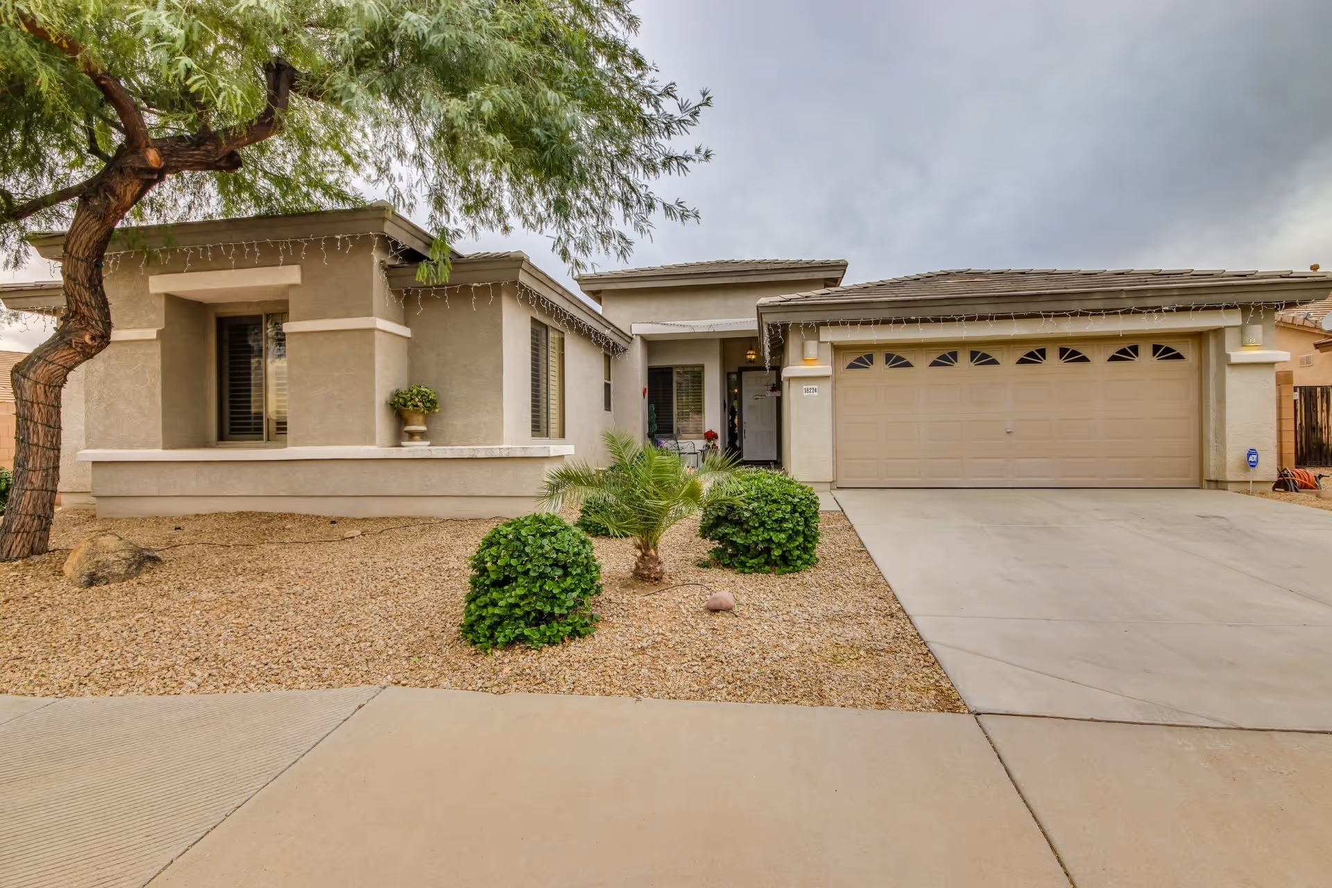 Front exterior view of a single-story residential building with a two-car garage, beige stucco walls, a small landscaped area with bushes and a tree, and a concrete driveway under a cloudy sky.