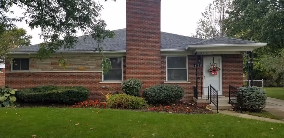 Single-story brick building with a central chimney, two windows, and a white front door decorated with a floral wreath. The building is surrounded by green grass, bushes, and trees.