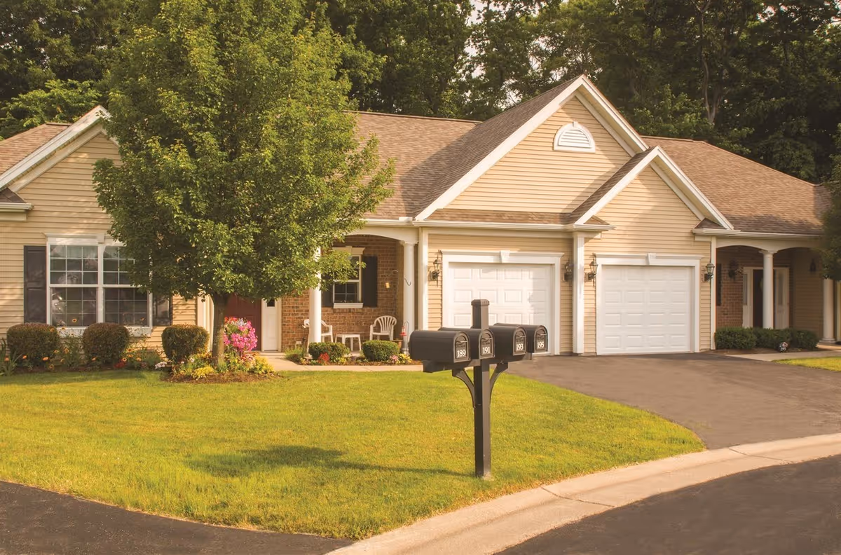 Exterior view of a residential building with beige siding and white garage doors, surrounded by green grass and trees. There are three black mailboxes on a post in the foreground and a paved driveway leading to the garages. The building has a porch area with white chairs and some landscaping with bushes and flowers.