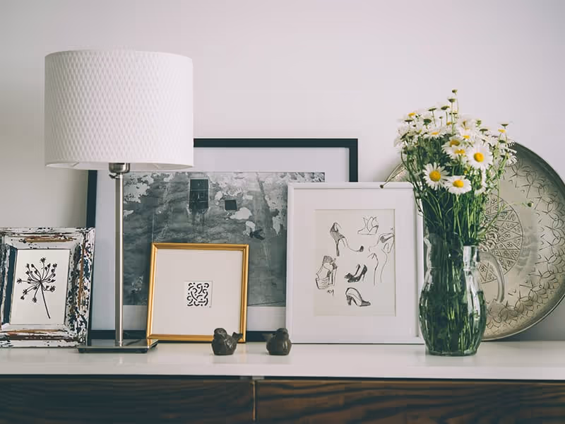 A white table or shelf with a white textured lamp, several framed pictures including a black and white photo, a drawing of shoes, and a decorative print, two small bird figurines, a clear glass vase with white daisies, and a large decorative metal tray leaning against the wall.
