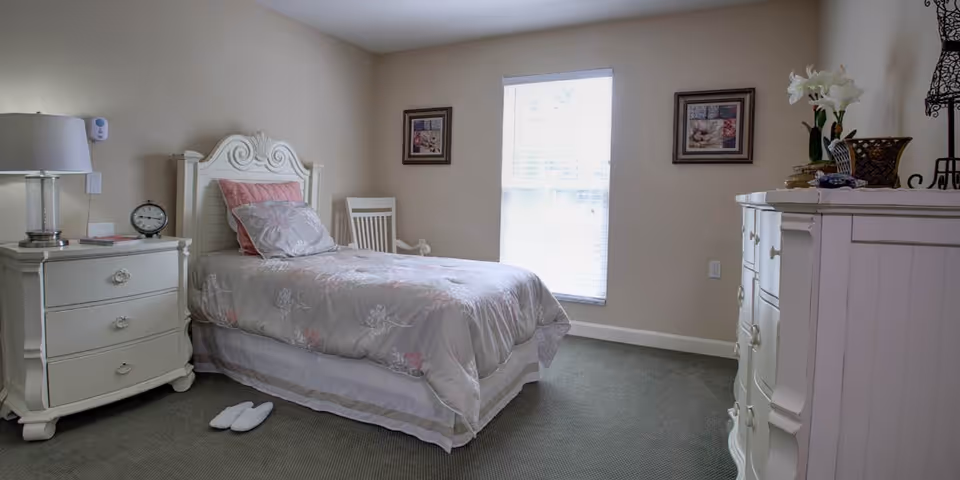 A neatly arranged bedroom with a single bed featuring a white ornate headboard, light-colored bedding with floral patterns, and two pillows. Next to the bed is a white nightstand with a lamp, an alarm clock, and some small items. A white wooden chair is placed against the wall near a window with blinds. On the right side, there is a white dresser with decorative items on top. The room has beige walls, carpeted floor, and two framed pictures hanging on the walls.