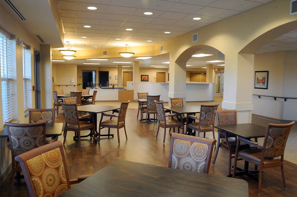 A spacious dining area in a senior living facility with multiple square tables and cushioned chairs featuring patterned upholstery. The room has wooden flooring, beige walls, large windows with blinds on the left side, and ceiling lights providing warm illumination. There are archways and a reception or service counter visible in the background.