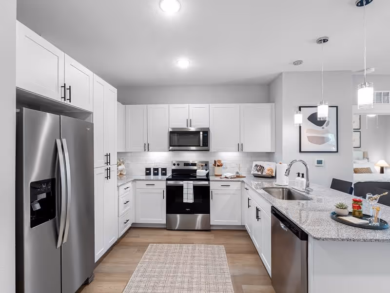 Modern kitchen with white cabinets, stainless steel refrigerator, oven, microwave, and dishwasher. Granite countertops with a sink and faucet, pendant lights hanging from the ceiling, and a small rug on the wooden floor. A framed abstract artwork is visible on the wall, and a glimpse of a bedroom can be seen in the background.