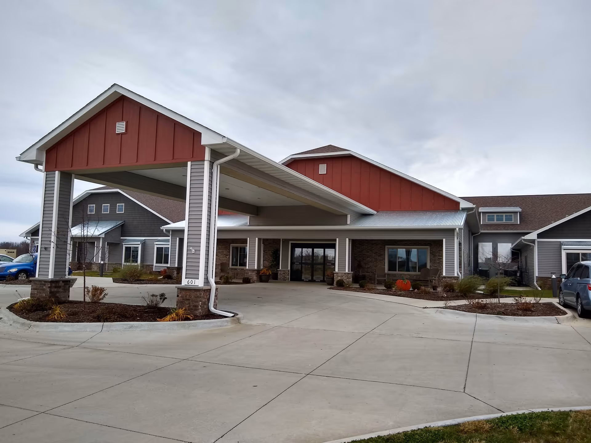 Exterior view of a senior living facility named Savannah Heights, showing the main entrance with a covered drop-off area, surrounded by a paved driveway and landscaped garden beds under a cloudy sky.