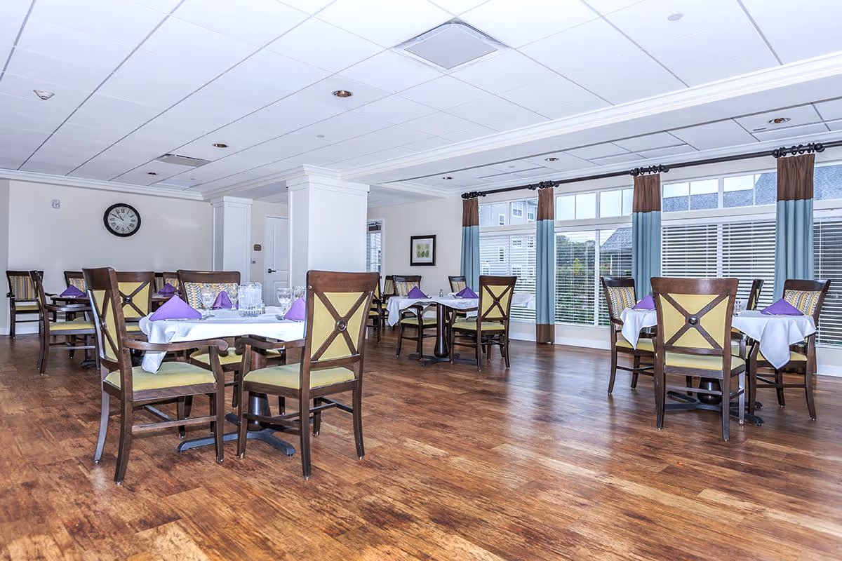 Bright dining room with multiple set tables and chairs on a wood floor and large windows with curtains.