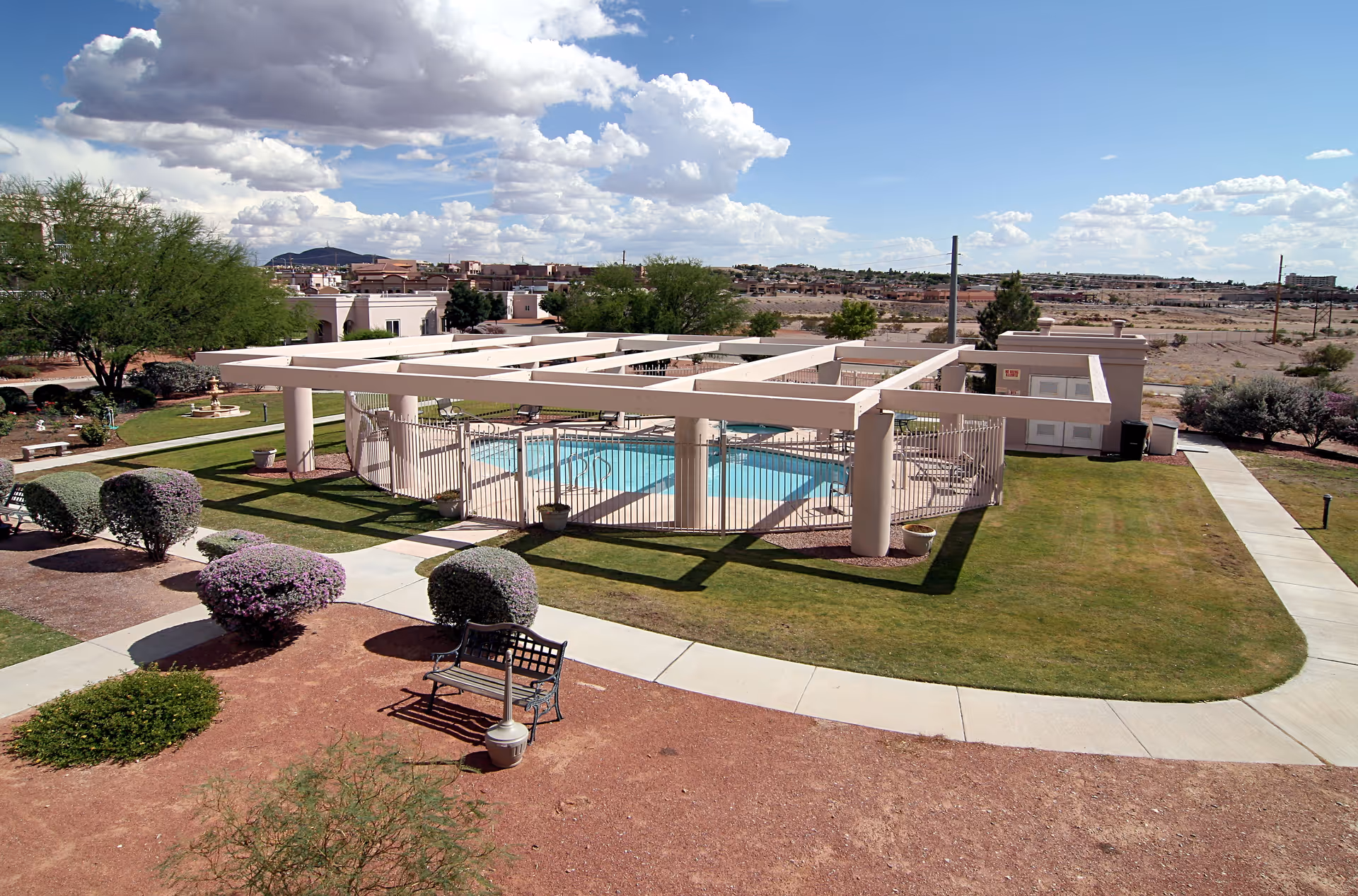 Outdoor view of a senior living facility's fenced swimming pool area with a pergola structure overhead. Surrounding the pool are green lawns, trimmed bushes, a bench, and paved walkways. The background shows a suburban neighborhood under a partly cloudy sky.