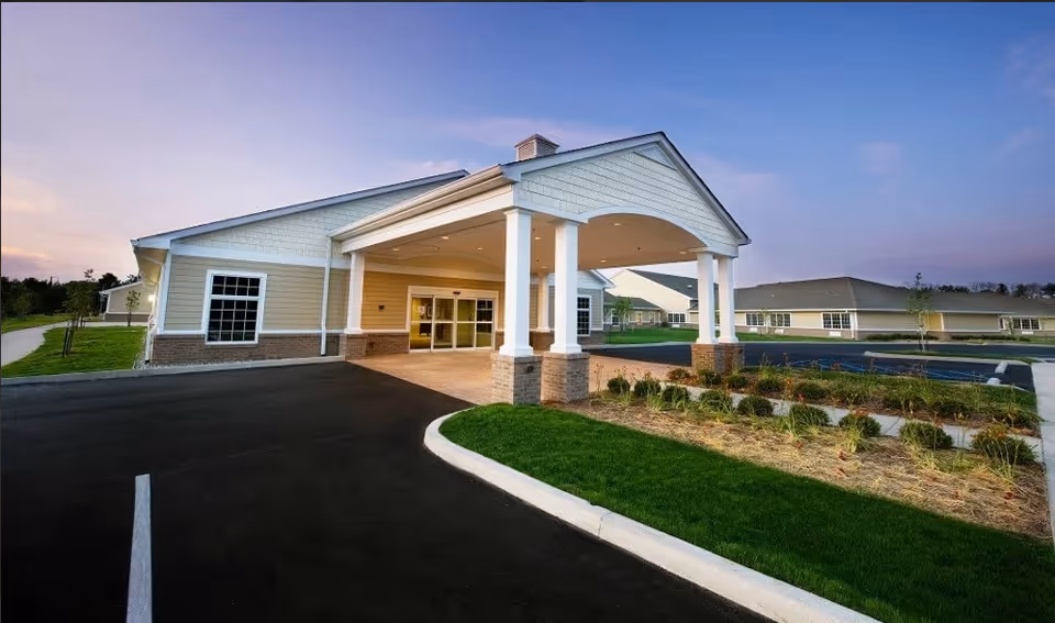 Front entrance of a single-story senior living facility with a covered porte-cochere, driveway, and landscaped lawns at dusk.