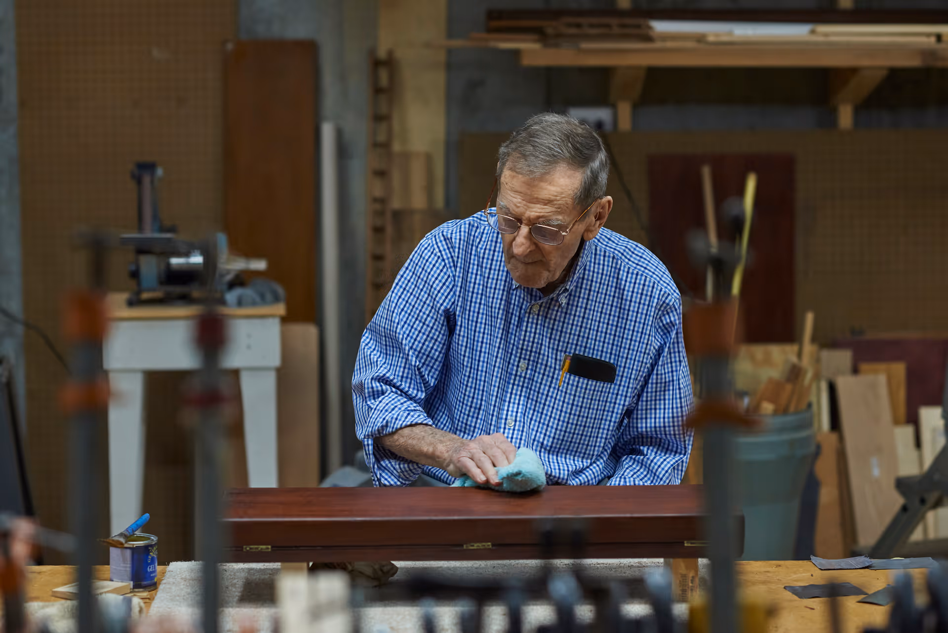 An elderly man wearing glasses and a blue checkered shirt is polishing a wooden piece in a workshop filled with woodworking tools and materials.