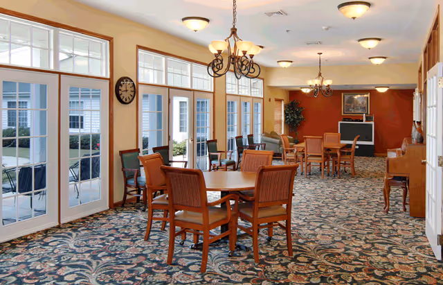 A spacious common area with multiple round wooden tables and chairs arranged on a patterned carpet. Large windows and glass doors line one side of the room, allowing natural light to fill the space. The room features warm lighting fixtures, a clock on the wall, a piano, a television, and some plants, creating a comfortable and inviting atmosphere.