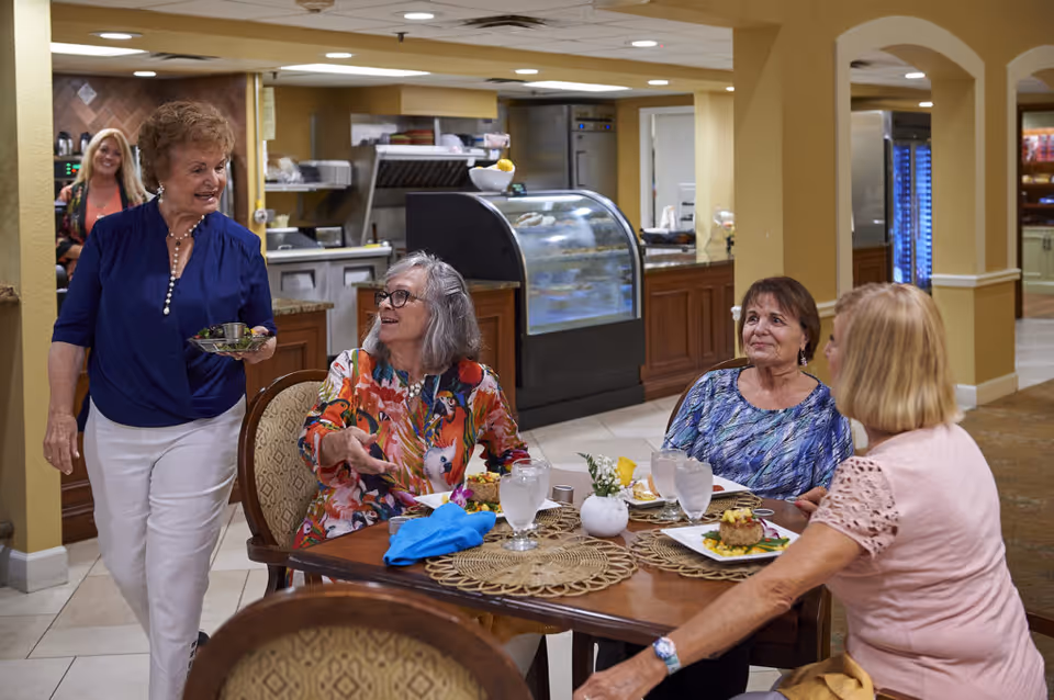 Four older women chat and share a meal at a dining table inside a communal dining area with a kitchen counter and display case in the background.