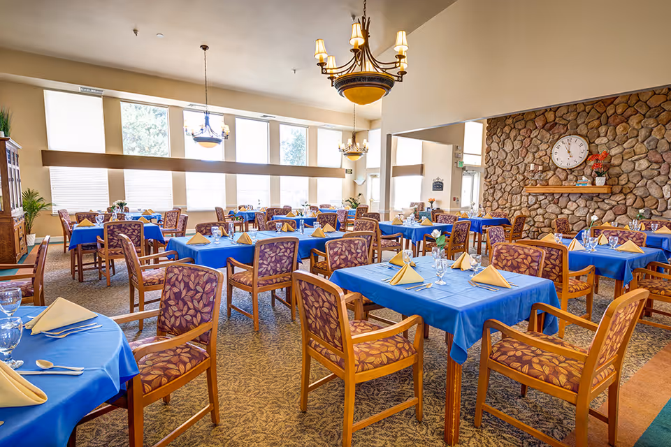 Bright dining room with multiple tables covered in blue tablecloths, set with napkins and glassware, and wooden chairs throughout.