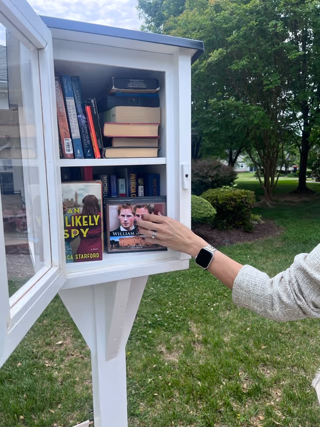 A person wearing a watch and a light-colored sweater is reaching into a small white outdoor book exchange box filled with books. The box is mounted on a white post and is located in a grassy area with trees and bushes in the background.