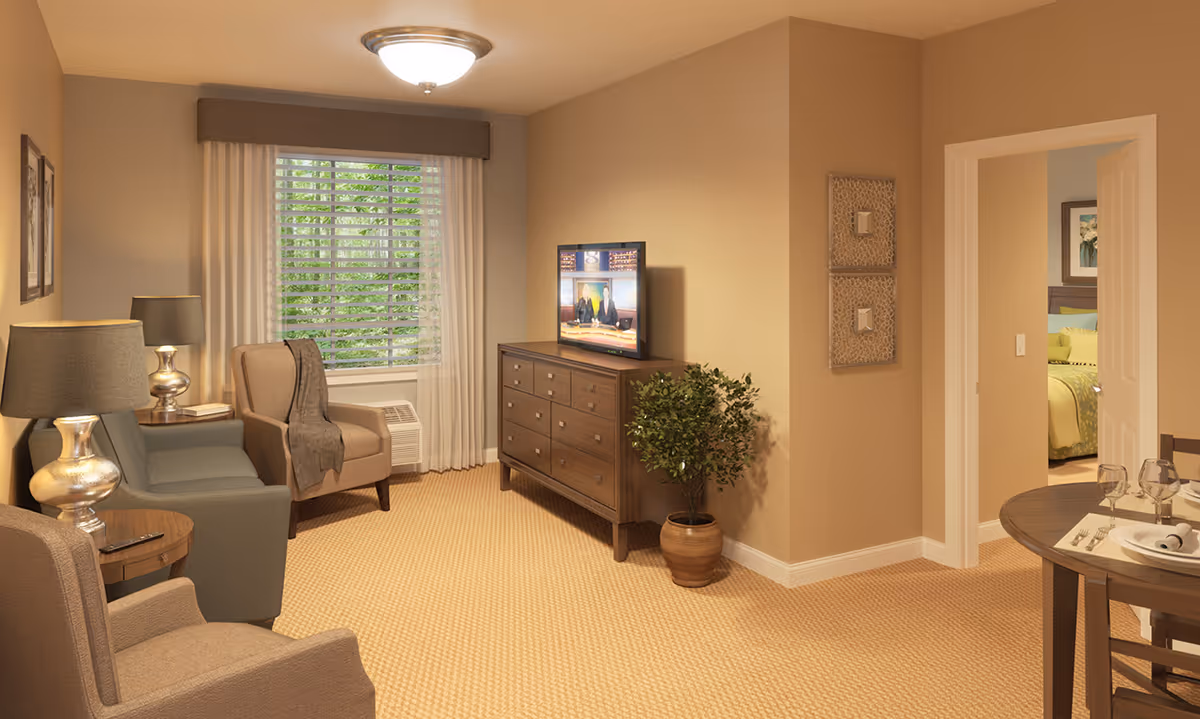 A cozy living room area with beige walls and carpet, featuring a window with white curtains and blinds. The room has a blue sofa, two beige armchairs, two silver table lamps, a wooden dresser with a flat-screen TV on top, and a potted plant beside the dresser. To the right, there is a round dining table set with plates, glasses, and silverware, and an open doorway leading to a bedroom with a bed and framed artwork on the wall.